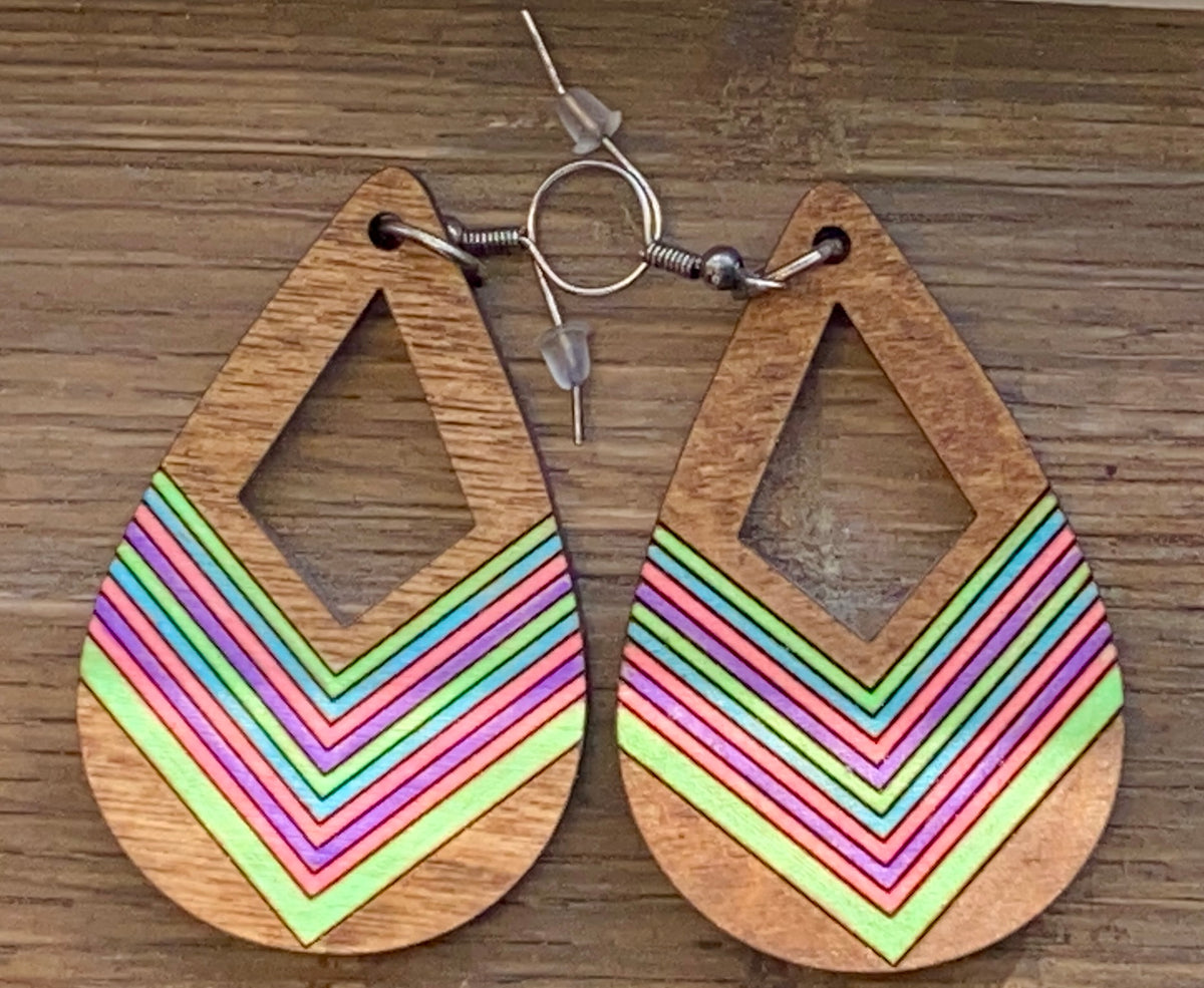 A pair of tear drop earrings with a geometric prairie style design, featuring layers of pink, green, and yellow stripes on a wooden texture.