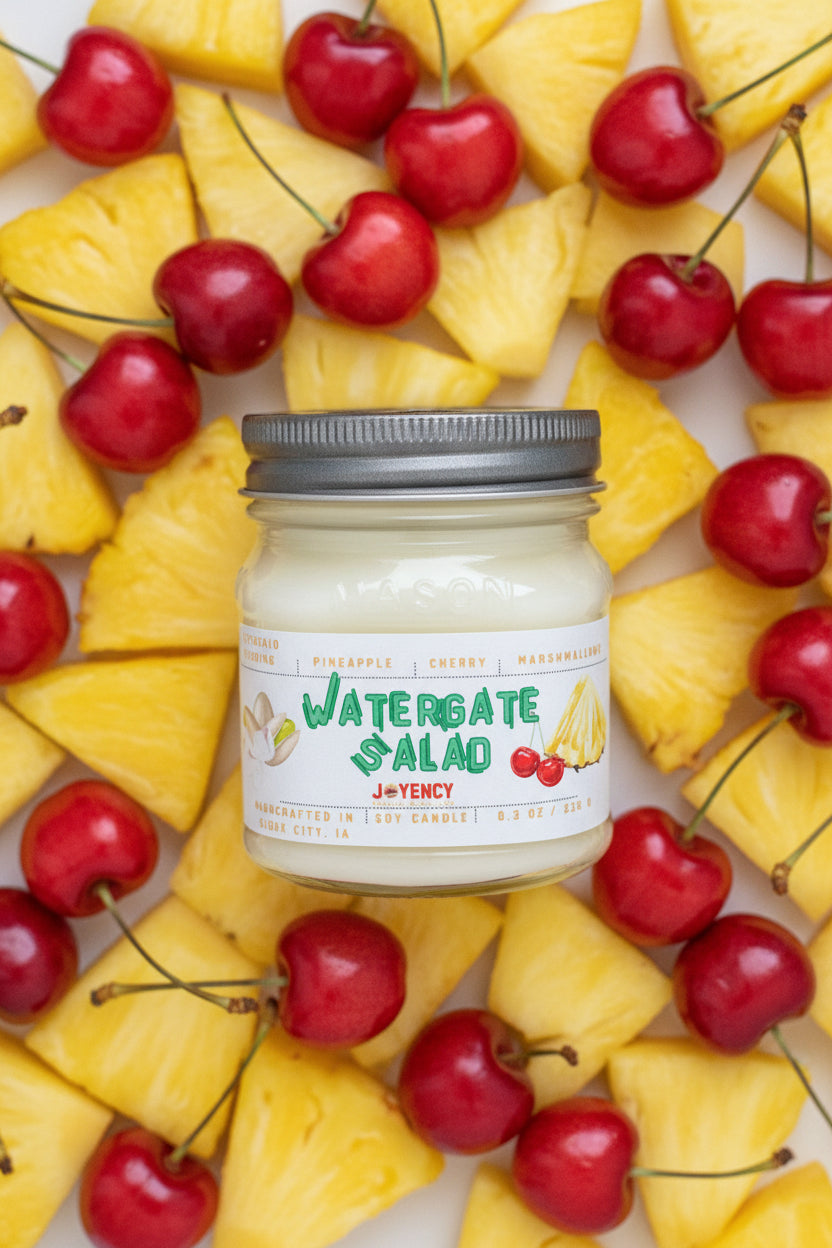 A jar candle labeled 'Watergate Salad' by J. of Yency, featuring a white wax fill, a silver metal lid with a floral cutout, and a white label with green and red text and illustrations.