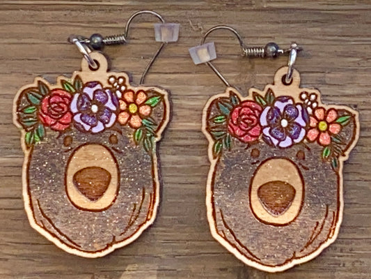 A pair of wooden earrings, each in the shape of a bear's head adorned with colorful floral patterns.