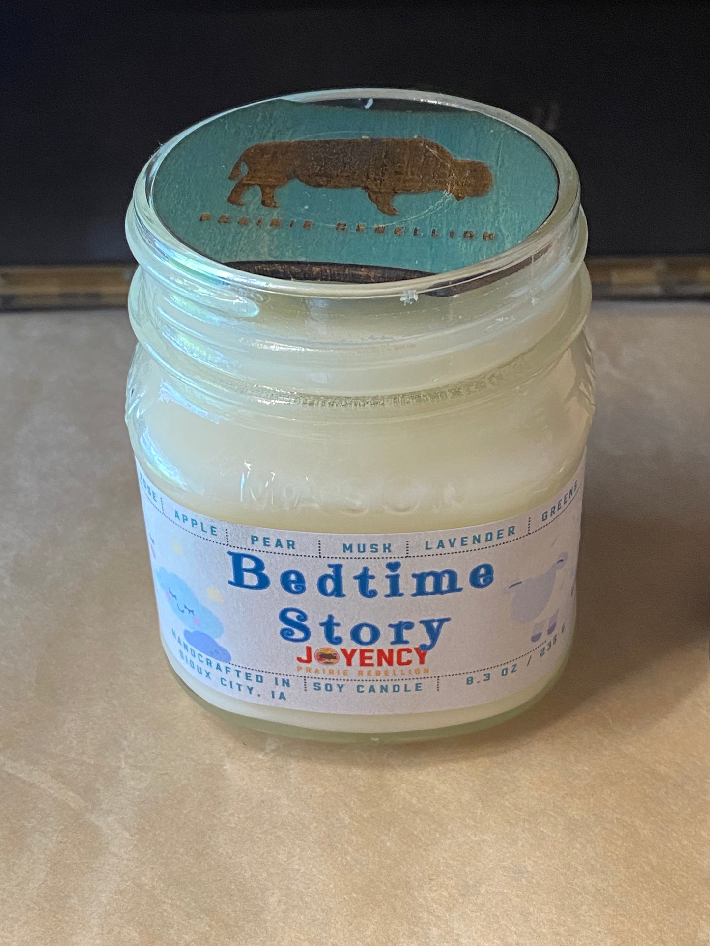 A glass jar candle with creamy off-white wax and a white label reading 'Bedtime Story' by Joyency. The lid has a turquoise label with a brown buffalo silhouette and the words 'Pioneer Wildflower.