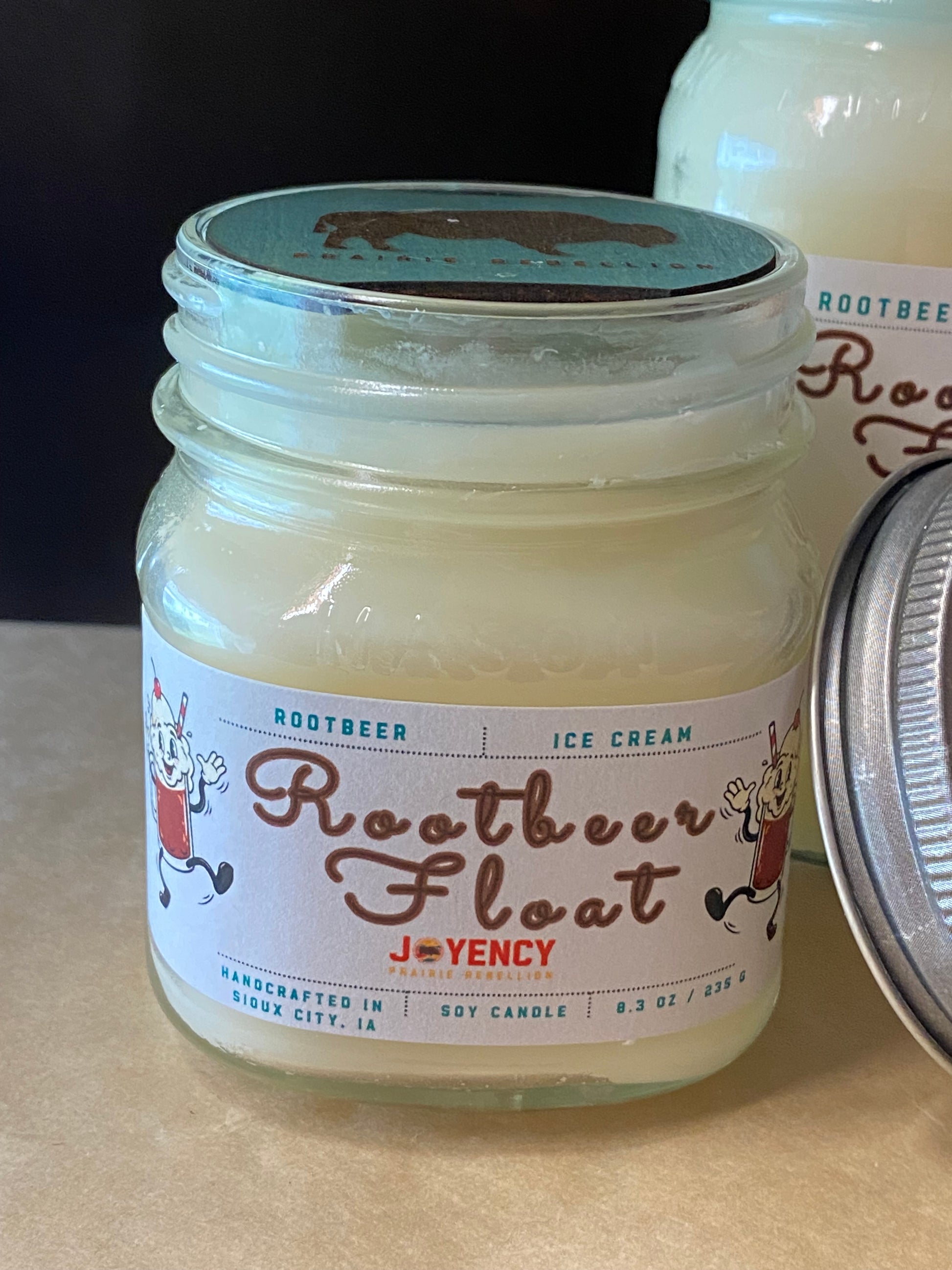 A glass mason jar candle labeled 'Rootbeer Float' by J*V*ENCY, filled with creamy off-white wax and shown with its metal lid beside it on a tabletop.
