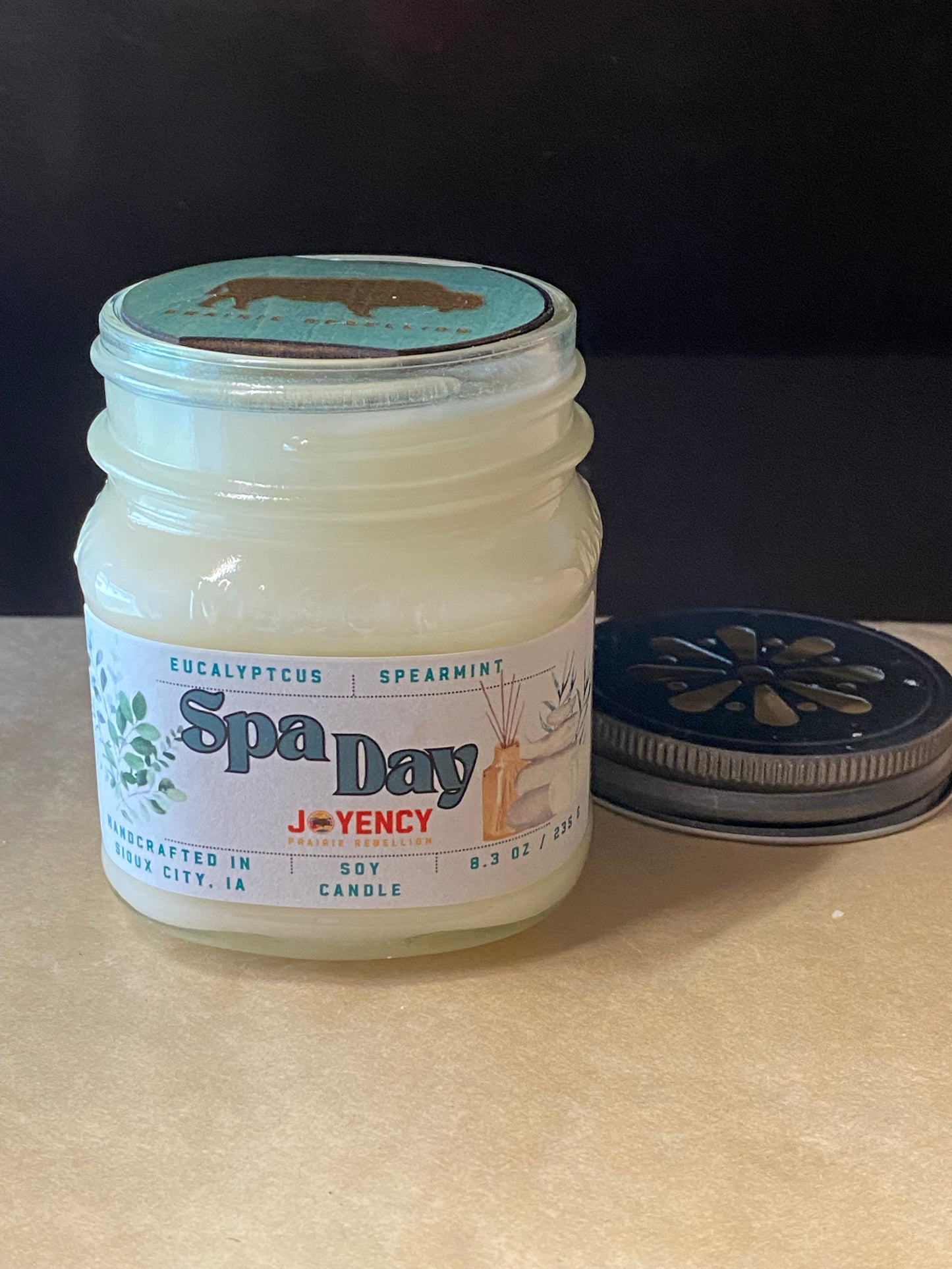 A glass jar candle labeled 'Spa Day' by Joyency, filled with pale creamy wax and topped with a turquoise lid, shown on a beige surface with its metal and turquoise lid placed beside it against a dark background.