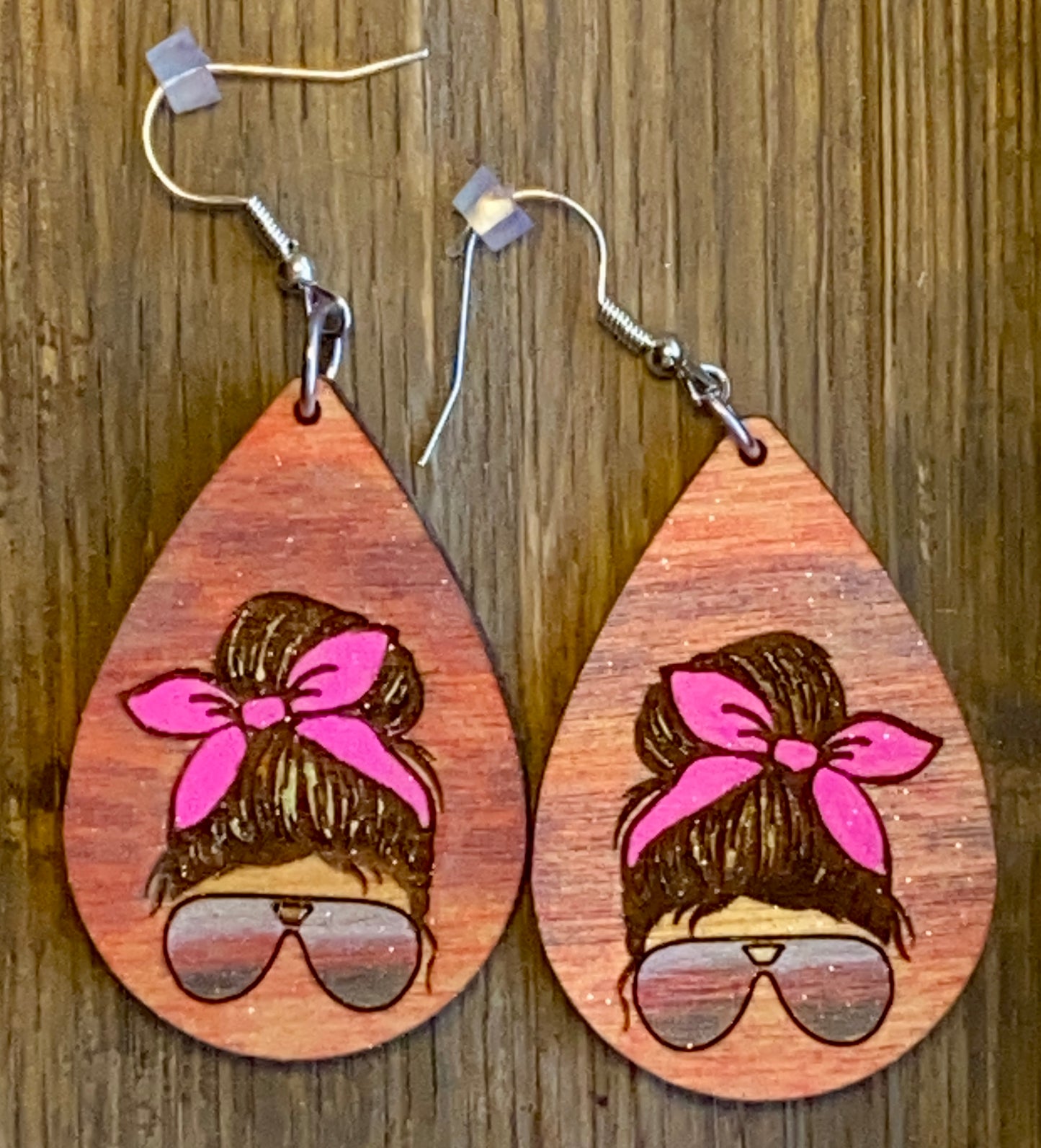 A pair of teardrop-shaped earrings with a design that includes sunglasses and a headband on a wooden surface.