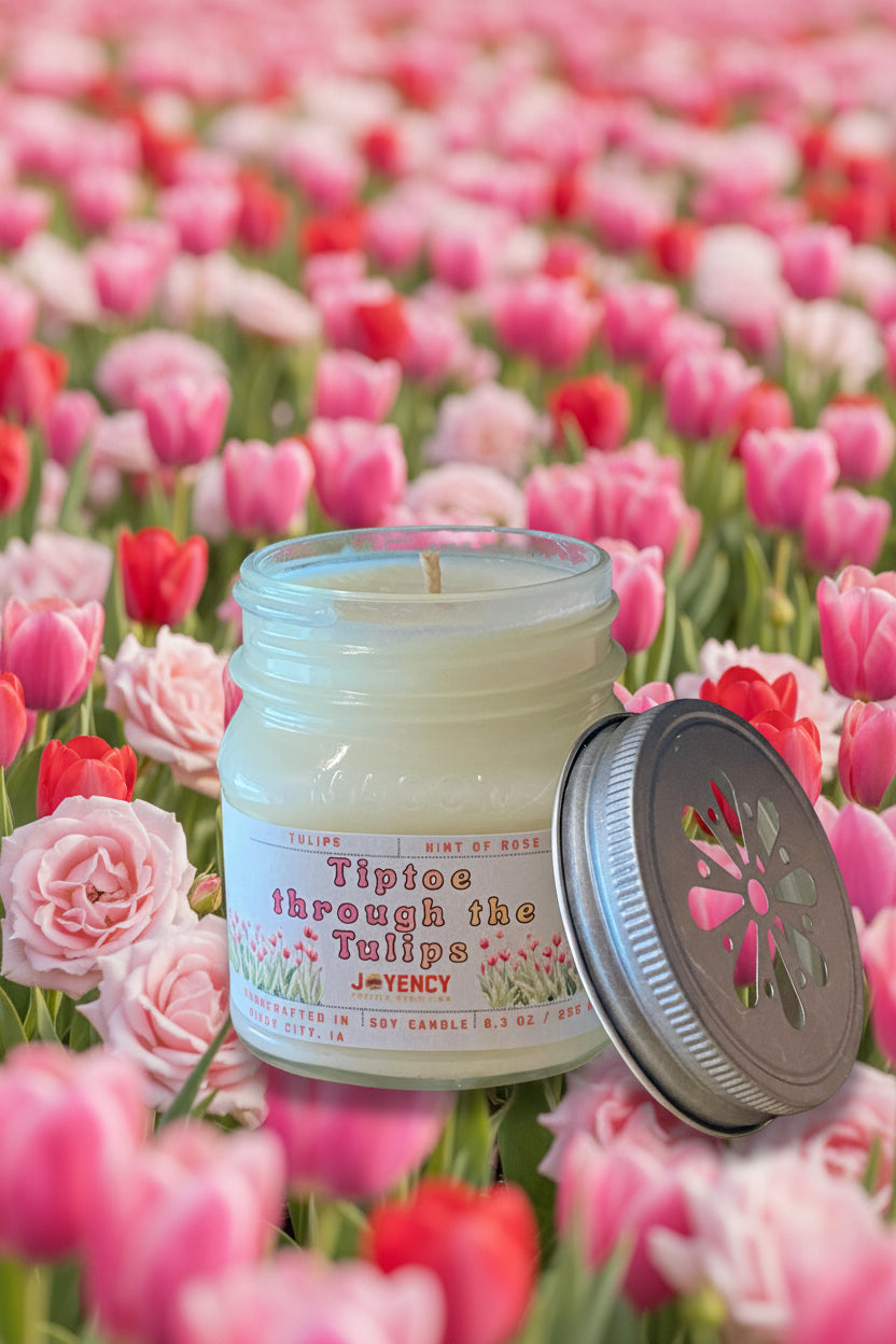 A glass jar candle with a white wax fill and a silver metal lid leaning against it on a beige surface. The label reads 'Tiptoe through the Tulips' by J.