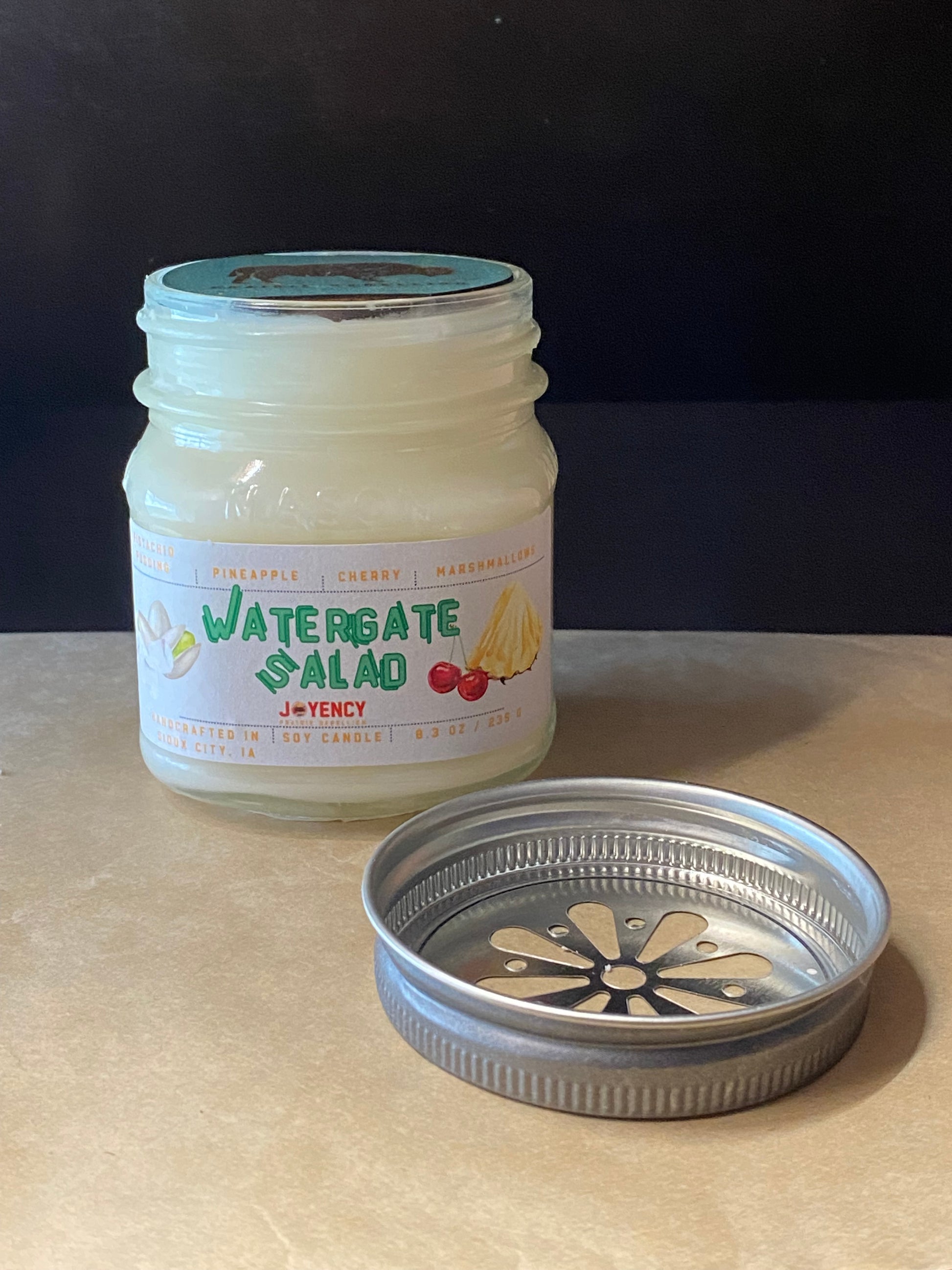 A glass jar candle labeled 'Watergate Salad' by Joyency, shown with its metal lid placed in front on a beige tabletop against a dark background.