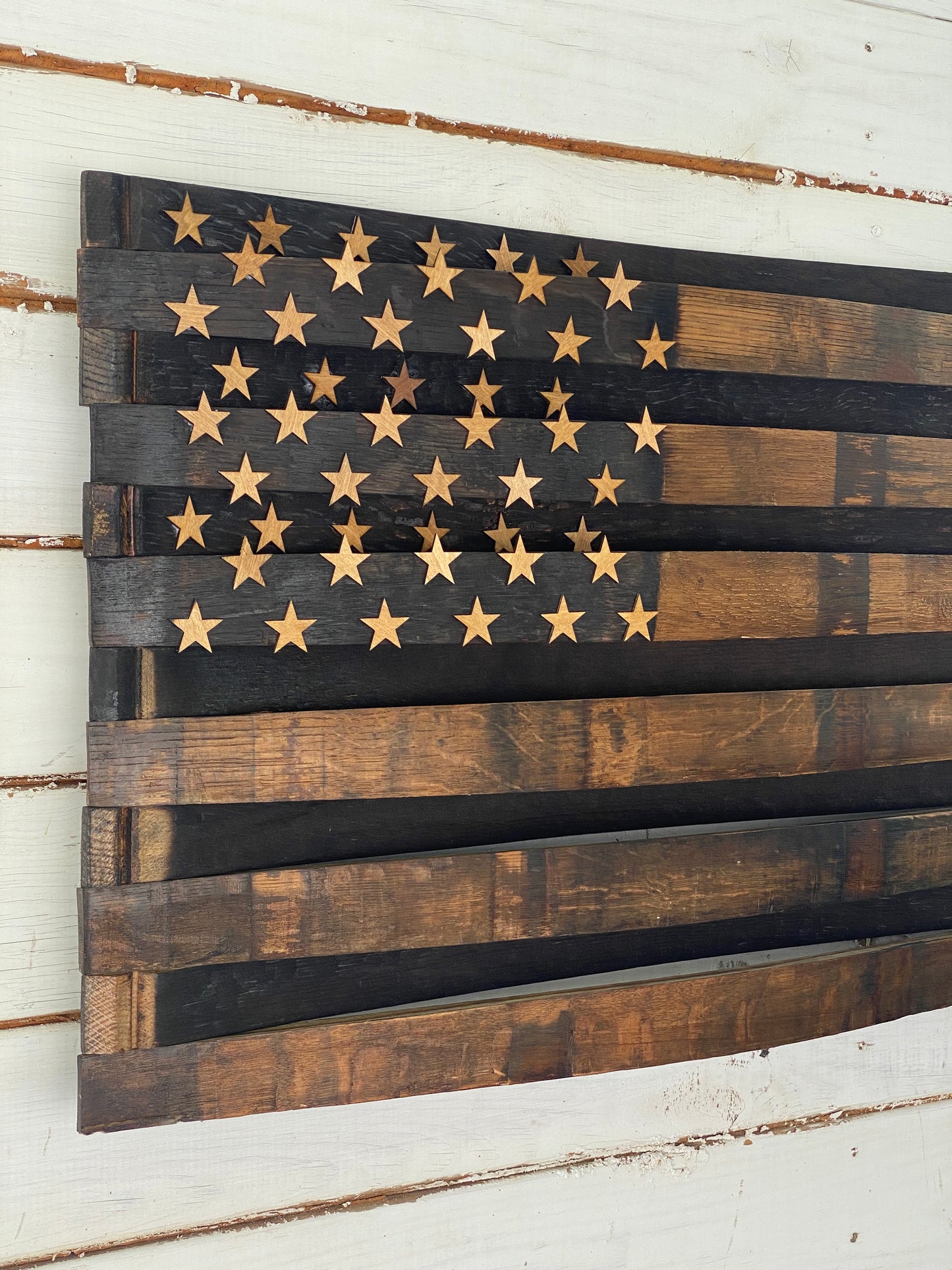 A rustic wooden American flag wall art piece made from dark and medium brown planks, with laser-cut gold stars on the left side, mounted on a white distressed wood plank wall.