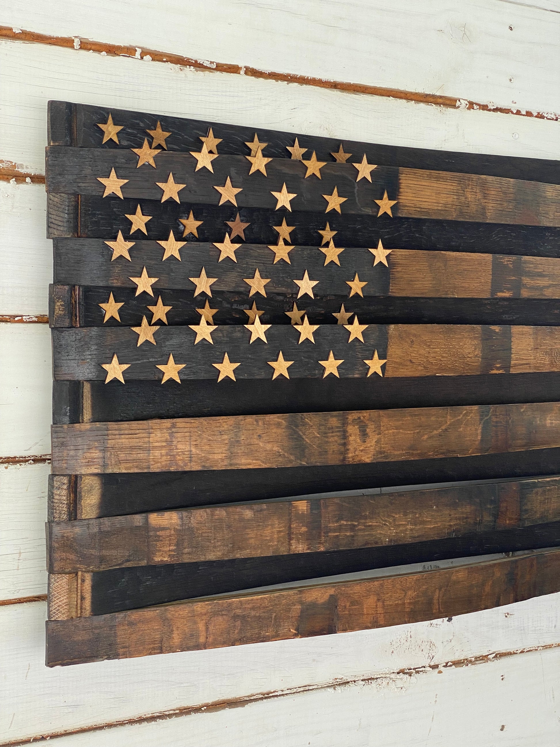 A rustic wooden American flag wall art piece made from dark and medium brown planks, with laser-cut gold stars on the left side, mounted on a white distressed wood plank wall.