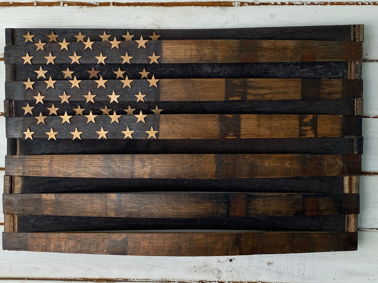 A rustic wooden American flag wall art piece made from stained wood planks, featuring a dark charred  field with natural wood-toned stars and alternating dark and medium brown stripes, mounted on a white distressed wood background.
