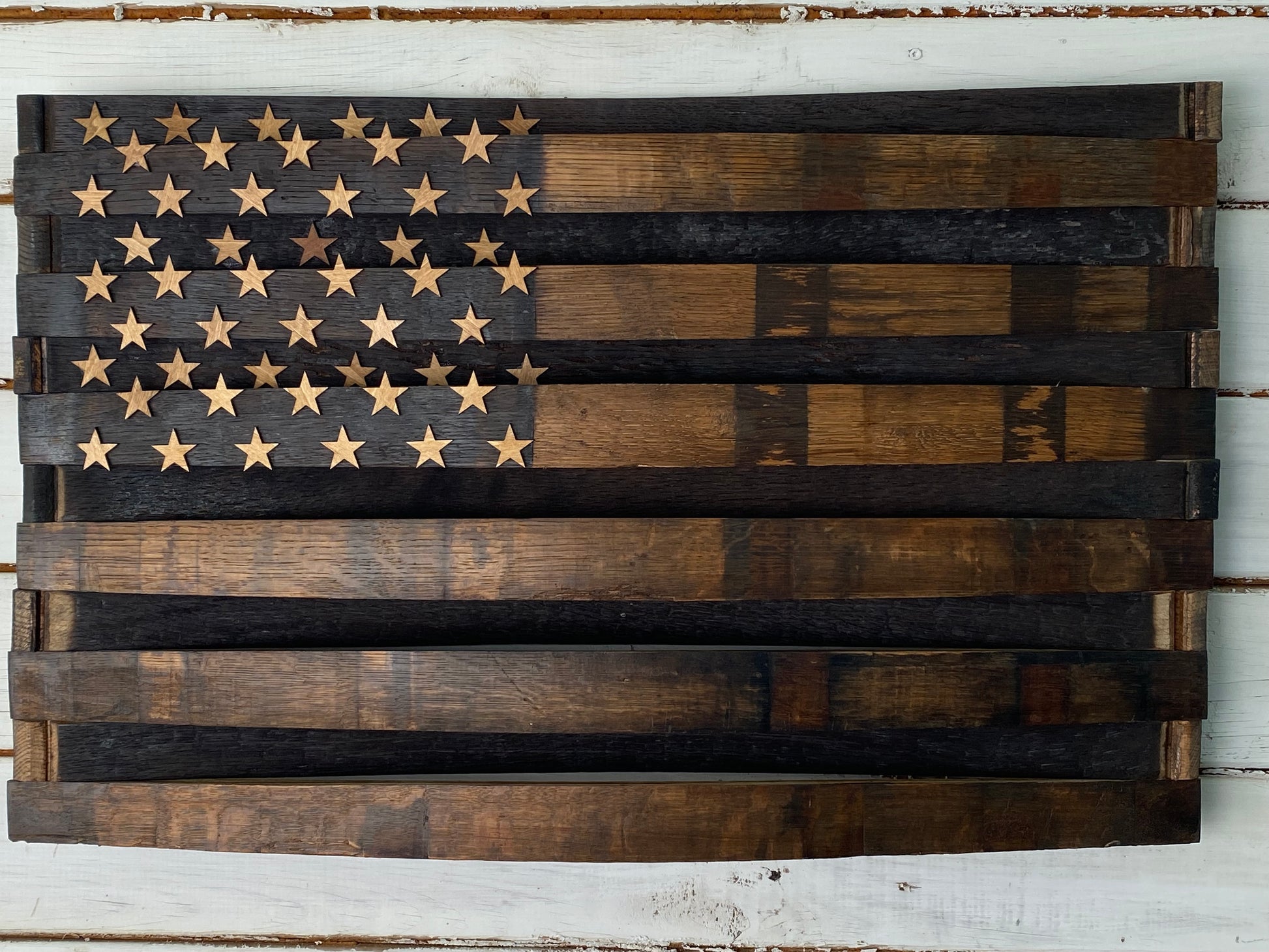A rustic wooden American flag wall art piece made from stained wood planks, featuring a dark charred  field with natural wood-toned stars and alternating dark and medium brown stripes, mounted on a white distressed wood background.