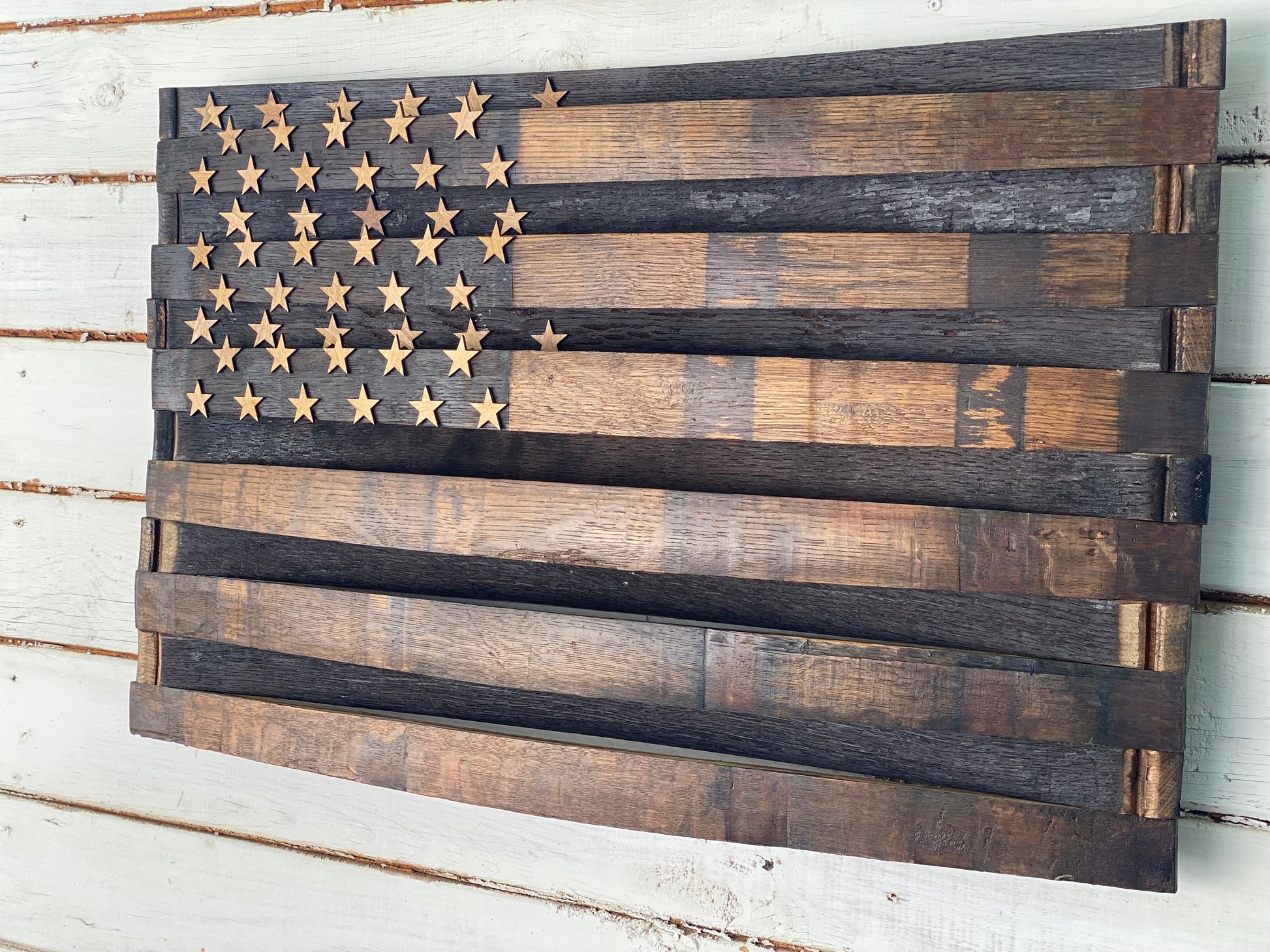 A rustic wooden American flag wall hanging made from stained wood planks, with a field of small star cutouts in the upper left, shown against a whitewashed wood plank wall.