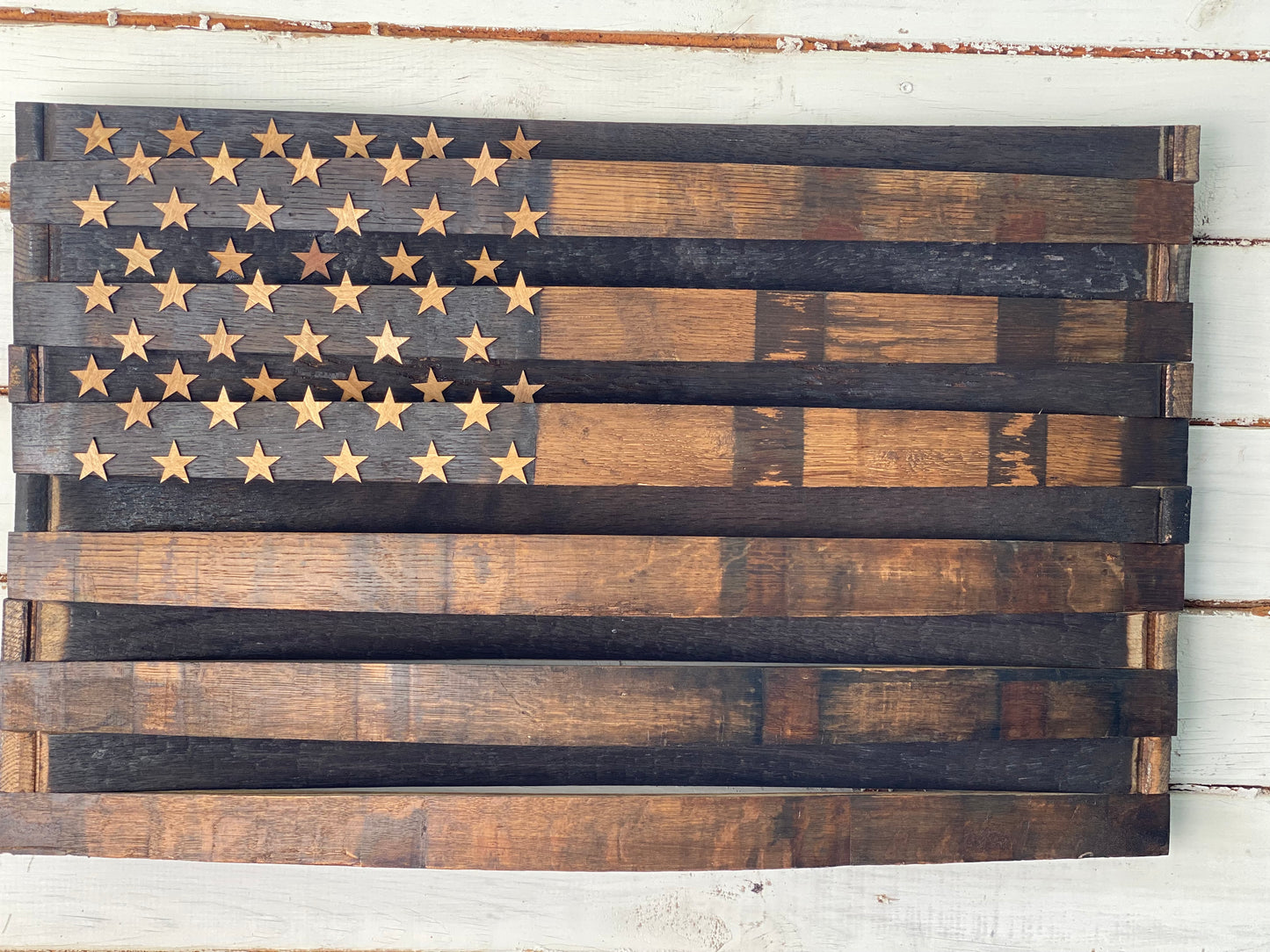 A rustic wooden American flag wall art piece made from stained wood planks, featuring a dark charred field with natural wood stars, shown against a white distressed wood background.