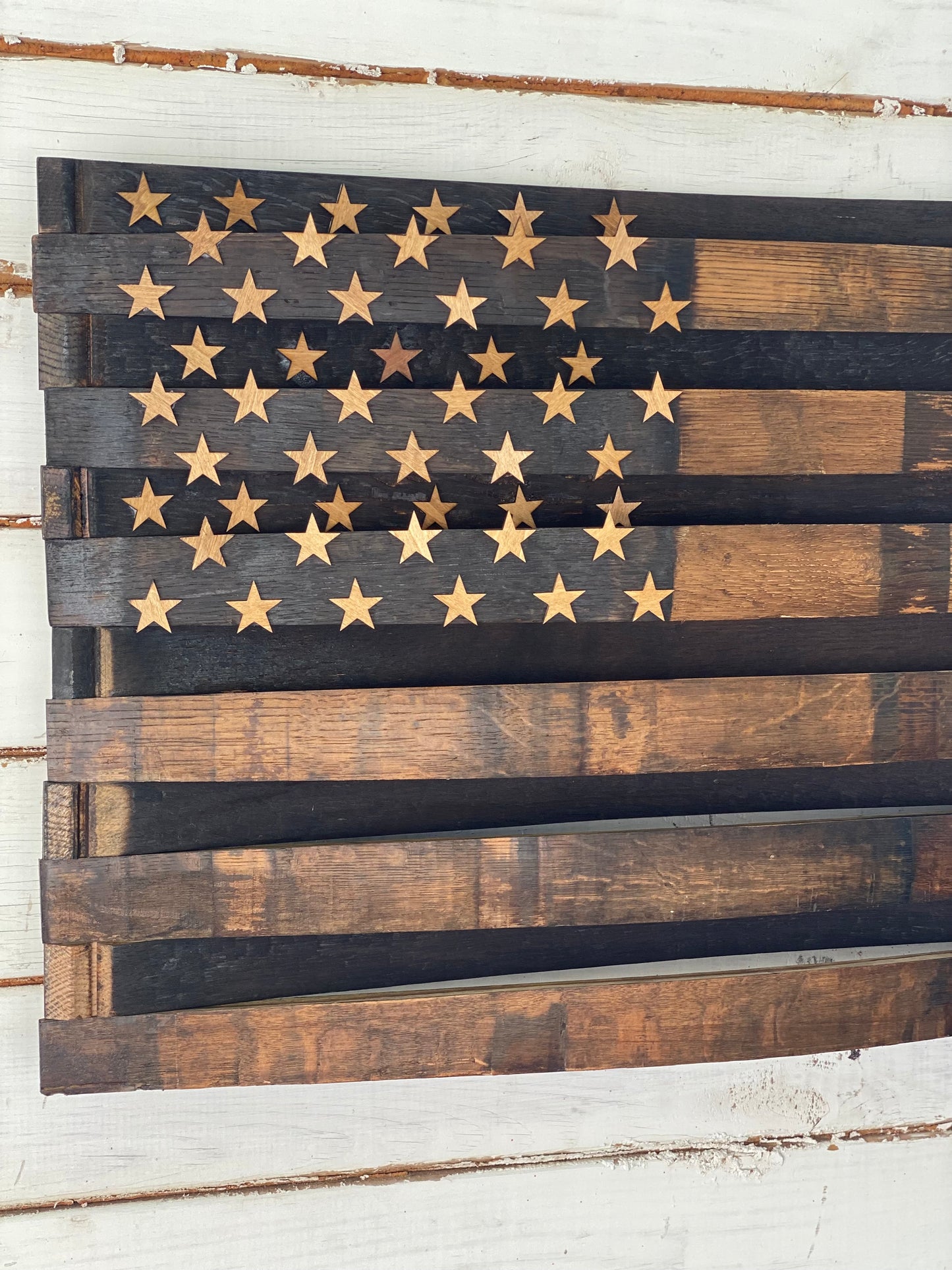 A rustic wooden American flag wall art piece made from dark and medium brown planks, with laser-cut gold stars on a dark field, shown against a white wood plank background.